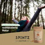 Frau steht mit ausgebreiteten Armen in einem Wald und schaut entspannt nach oben; im Vordergrund ein Produktbild von „SPINTZ Reishi-Kapseln“ mit dem Schriftzug „Supplements: wohldosierte Natur“
